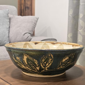 May include: A handcrafted ceramic bowl with a dark teal exterior and a light beige interior. The bowl features a leaf design in a lighter tan color. The bowl is sitting on a wooden surface.