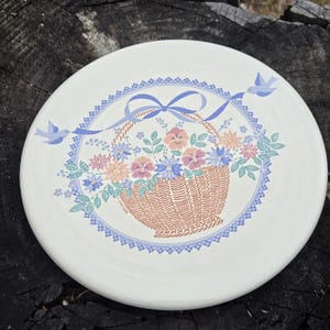 May include: Round, white ceramic plate with a floral basket design. The basket is filled with pink, blue, and peach flowers. A blue ribbon and two blue birds decorate the border. The plate is approximately 9 inches in diameter.