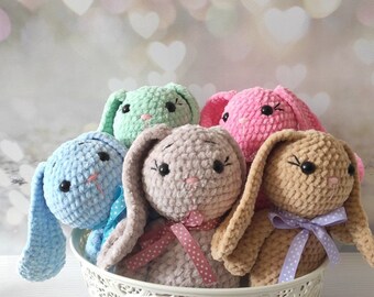 Könnte beinhalten: Ein dekorativer weißer Korb gefüllt mit sechs gehäkelten Hasen in Pastelltönen. Die Häschen sind blau, grün, rosa und beige, jedes mit langen Ohren, einer Schleife und Knopfaugen. Der Korb steht auf einer weißen Holzoberfläche.