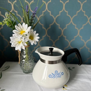 May include: White teapot with a blue floral design and a black handle. The teapot has a silver band around the middle and a silver lid. The teapot is sitting on a white tablecloth with a floral pattern.