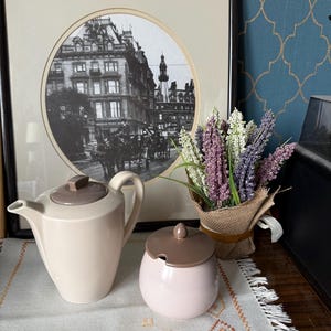 May include: A framed monochrome print of a city scene, a cream teapot with a brown lid, and a pink sugar bowl with a brown lid. A small bouquet of lavender and white flowers in a burlap wrap sits to the right.