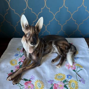 May include: A brown and white ceramic figurine of a German Shepherd dog lying down with its tongue sticking out. The figurine is on a white tablecloth with embroidered flowers.