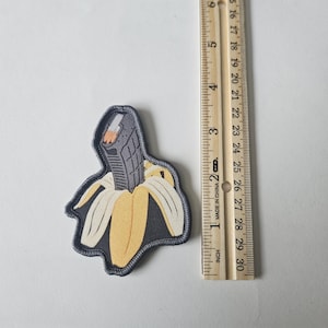 Op de afbeelding: Een patch met een banaan met een pistool erin. De patch heeft een zwarte rand en een gele en witte bananenschil. Het pistool is grijs en zwart. De patch is ongeveer 7,6 cm lang.