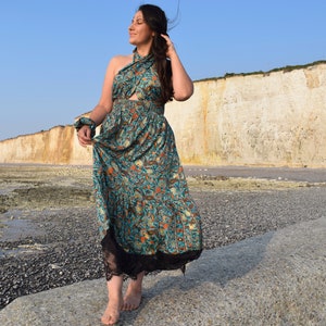 May include: A woman wearing a blue and gold patterned maxi dress with a black lace trim. The dress has a halter neckline and a fitted bodice. She is standing on a rocky beach with a white cliff in the background.