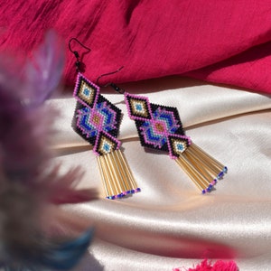 May include: A pair of geometric beaded earrings with a pink, blue and gold colour scheme. The earrings feature a diamond-shaped design with a tassel of gold beads.