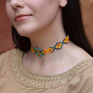 Peut inclure: Un collier ras du cou en perles avec un motif géométrique. Le collier présente un motif de perles jaunes, noires et turquoise. Le collier est porté autour du cou.