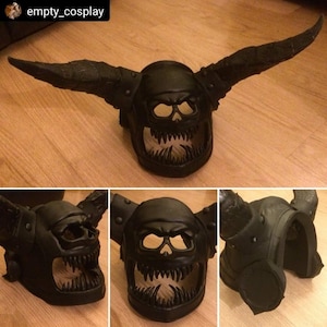 Pu&ograve; includere: Un casco di plastica nero con corna e un viso a forma di teschio. Il casco ha un design dettagliato con punte e un ghigno pieno di denti.