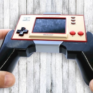 May include: A handheld Game & Watch console with a cream-coloured face, black screen, red buttons, and a black directional pad. The console is held by black handles. The text "GAME & WATCH" and "SUPER MARIO BROS." are visible.