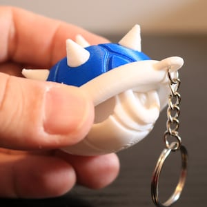May include: A blue and white 3D printed keychain shaped like a spiky shell. The keychain has a silver chain and ring.