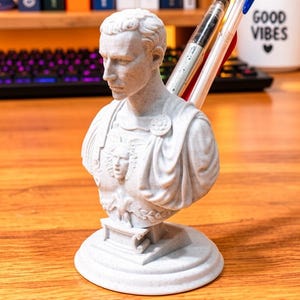 May include: A light gray, stone-textured bust of a Roman figure serving as a desk organizer. The sculpture features a detailed chest plate with a Gorgon head emblem and includes two cylindrical openings at the back for holding pens or pencils.