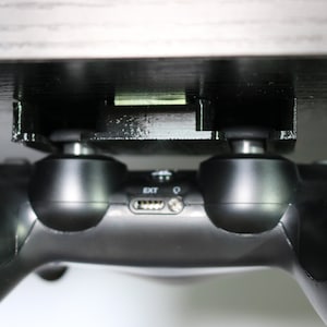 May include: A black video game controller is mounted to a black plastic holder that is attached to a dark wood surface. The controller is hanging upside down.