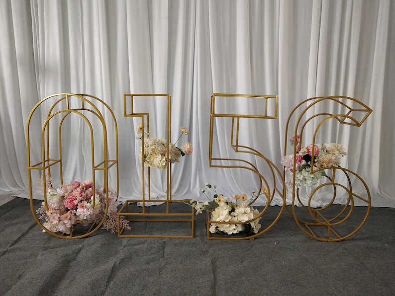 May include: Gold metal number frames with floral arrangements. The numbers are 0, 1, 5, and 6. The flowers are pink, white, and cream.