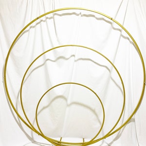 May include: A set of three gold metal hoops, arranged in a nested circle design, with a base stand. The hoops are used for wedding or event decor.