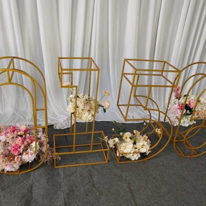 May include: Gold metal frame numbers 1, 5, and 6 decorated with pink and white flowers. The numbers are freestanding and can be used for a wedding, party, or other event.