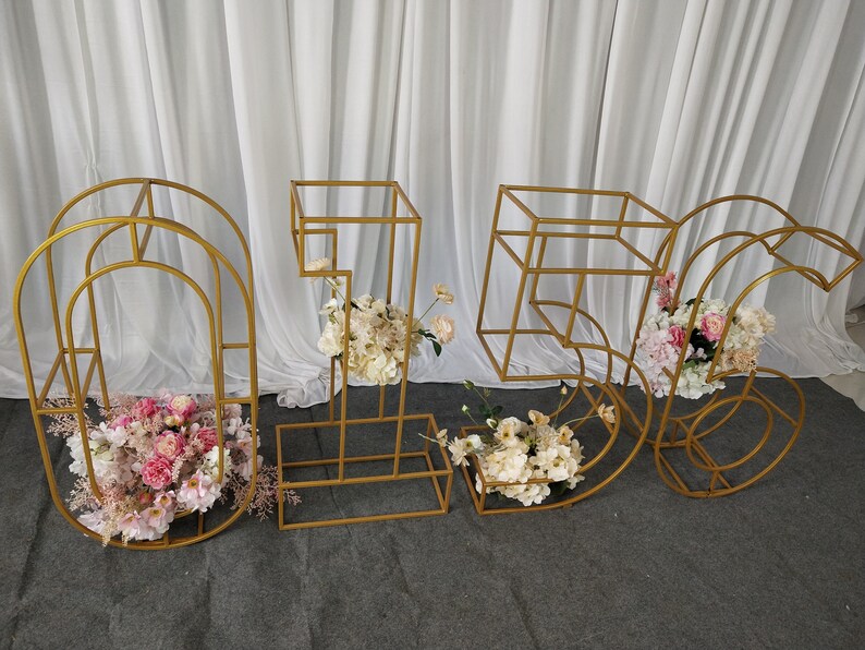 May include: Gold metal frame numbers 1, 5, and 6 decorated with pink and white flowers. The numbers are freestanding and can be used for a wedding, party, or other event.