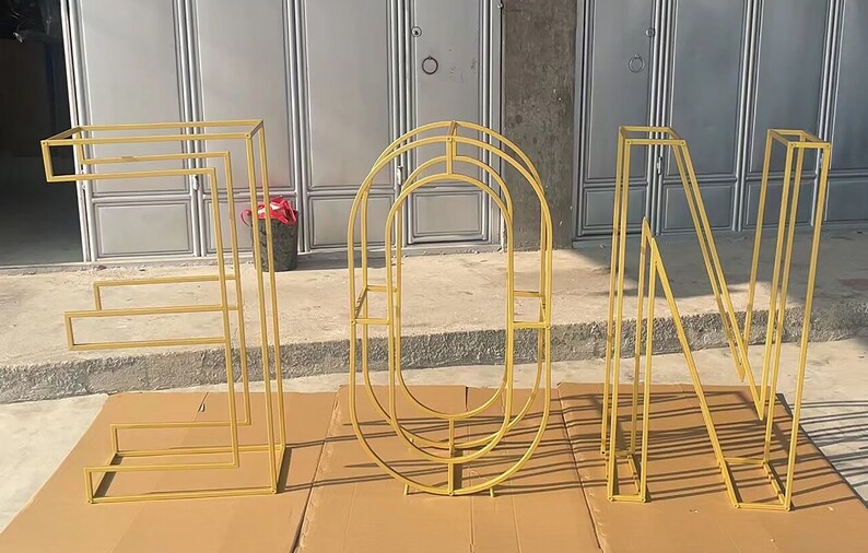 May include: Four gold metal letters spelling out the word "EON" standing on a brown surface. Each letter is made of a series of connected metal bars.