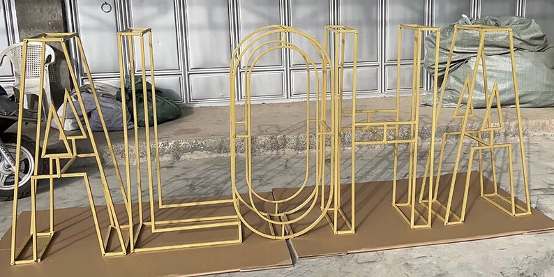 May include: Gold metal letters spelling out the word "ALOHA" in a modern, geometric style. The letters are freestanding and appear to be made of thin metal rods.