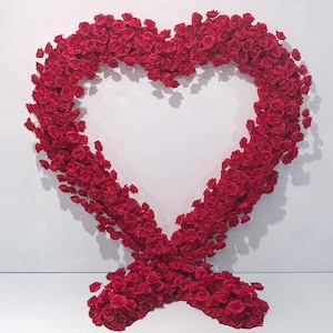 Könnte beinhalten: Eine große, herzförmige Blumengesteck aus vielen roten Rosen. Das Herz ist in der Mitte offen und steht vor einem weißen Hintergrund. Die Rosen sind dicht gepackt, wodurch ein voller, strukturierter Eindruck entsteht.