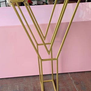 May include: A gold metal stand with a geometric design. The stand has a V-shape with multiple bars forming a rectangular base.