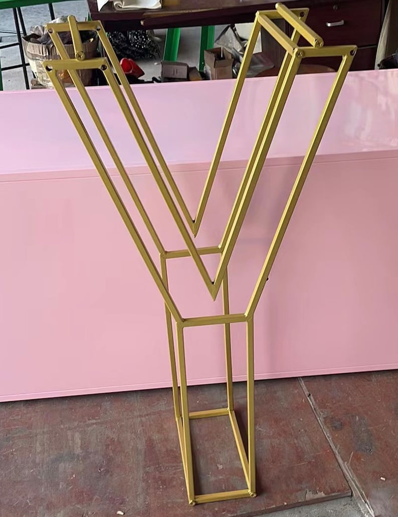 May include: A gold metal stand with a geometric design. The stand has a V-shape with multiple bars forming a rectangular base.