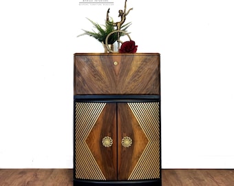 May include: A vintage wooden cabinet with a black and gold geometric design. The cabinet has a single drawer and two doors with gold handles. The cabinet is sitting on a wooden floor.