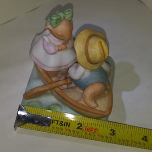 May include: A ceramic figurine of two rabbits in a boat. The rabbit in the front is wearing a straw hat and the rabbit in the back is wearing a green bow. The boat is brown and has a green and blue base.