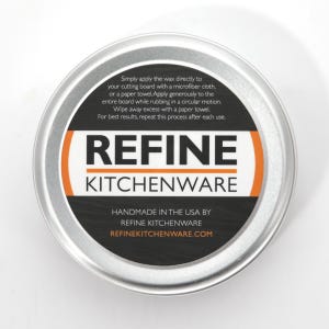 May include: A circular, silver metal tin containing Refine Kitchenware cutting board wax. The black label features white and orange text, including usage instructions and the website refinekitchenware.com. Made in the USA.