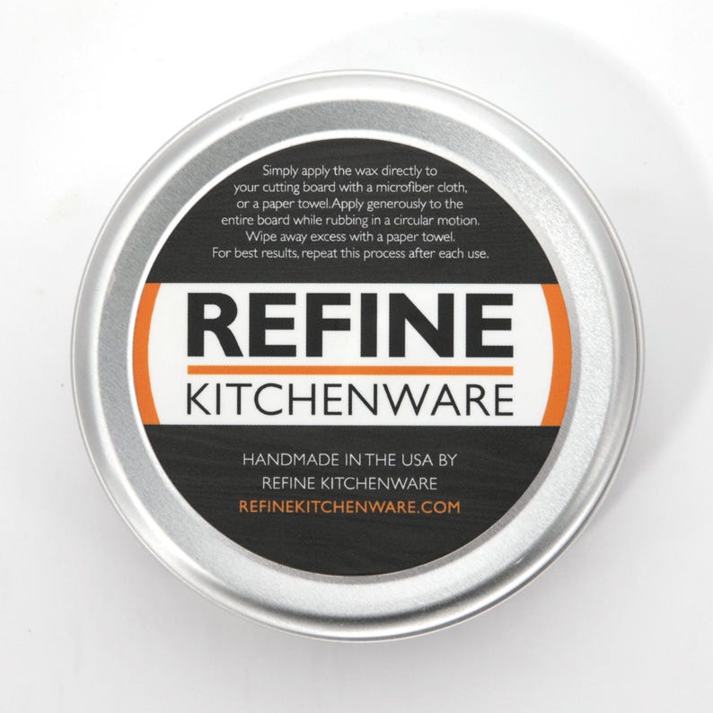 May include: A circular, silver metal tin containing Refine Kitchenware cutting board wax. The black label features white and orange text, including usage instructions and the website refinekitchenware.com. Made in the USA.