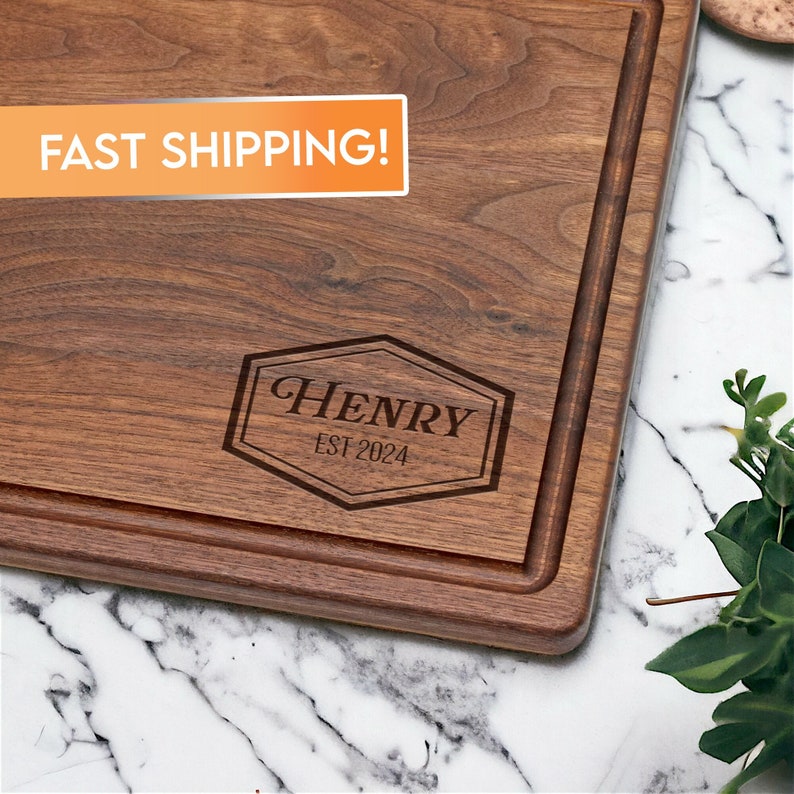 May include: A brown wooden cutting board with a personalized inscription that reads "HENRY EST 2024" in a hexagonal frame.