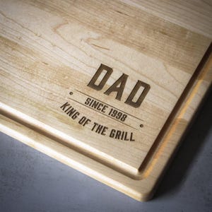 A wooden cutting board with the text "DAD SINCE 1998 KING OF THE GRILL" engraved on it.