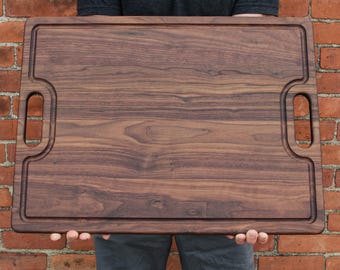 May include: A rectangular dark brown wooden cutting board with a visible natural wood grain pattern. The board features a recessed juice groove running along the perimeter and two integrated cutout handles on the left and right sides.