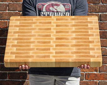 May include: A rectangular end-grain wooden cutting board featuring a light-colored, natural wood finish with a repeating brick-like pattern of joined wood blocks. The board has a thick, solid construction with smooth, rounded edges and corners.