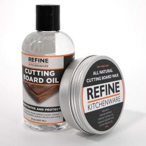 May include: A clear bottle of cutting board oil and a silver tin of cutting board wax. The oil bottle has a black cap and a label that reads "REFINE KITCHENWARE CUTTING BOARD OIL". The wax tin has a black and white label that reads "ALL NATURAL CUTTING BOARD WAX".