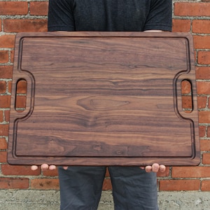 A person is holding a large wooden cutting board in front of a brick wall.