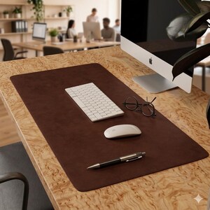 Könnte beinhalten: Eine braune, rechteckige Schreibtischunterlage mit einer weißen Tastatur, Maus und Stift. Die Unterlage liegt auf einem Holzschreibtisch mit einem Computermonitor und einer Brille. Der Hintergrund zeigt eine Büroumgebung.