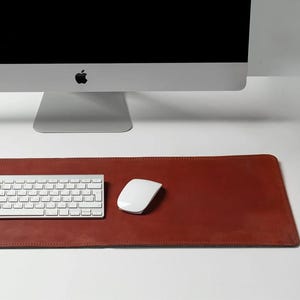 May include: A red-brown leather desk mat with a white keyboard and mouse. A silver computer monitor with the Apple logo is in the background. The desk mat is rectangular and has a stitched border.