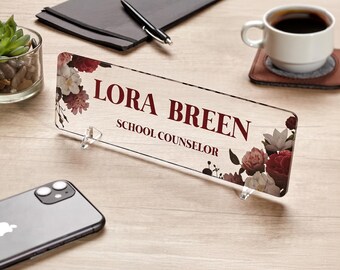 May include: A clear acrylic desk nameplate with a floral design and the text "Lora Breen School Counselor" in burgundy lettering.