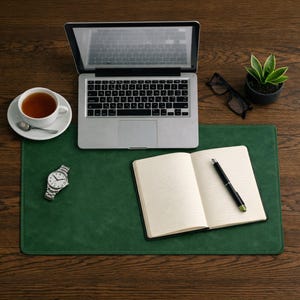 Könnte beinhalten: Eine grüne Schreibtischunterlage mit Laptop, offenem Notizbuch, Stift, Uhr, Brille, einer Tasse Tee und einer kleinen Topfpflanze auf einem Holzschreibtisch.