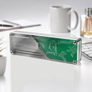 May include: A clear acrylic desk nameplate with a silver background and green circuit board design. The nameplate reads "Adam Johnson, System Administrator".