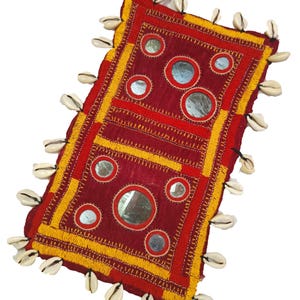 Può includere: Tessuto rettangolare fatto a mano, rosso e giallo, ornato con piccoli specchi rotondi e bordato con conchiglie bianche. Il design presenta motivi ricamati e un bordo con frange, creando un pezzo decorativo unico.