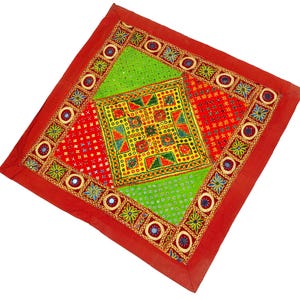 May include: A red square cushion with a colorful patchwork design. The cushion is decorated with embroidered flowers, geometric patterns, and small mirrors. The fabric is a mix of green, yellow, red, and blue.