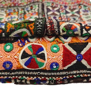 K&ouml;nnte beinhalten: Nahaufnahme eines farbenfroh bestickten Textils. Das Design zeigt geometrische Muster, darunter rote, wei&szlig;e, schwarze und orangefarbene Formen, mit Akzenten in Gr&uuml;n, Blau und Gelb. Das Textil scheint Teil eines gr&ouml;&szlig;eren St&uuml;cks zu sein, m&ouml;glicherweise einer Tasche oder eines Kleidungsst&uuml;cks.
