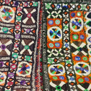K&ouml;nnte beinhalten: Nahaufnahme eines farbenfrohen Textils mit aufwendiger Stickerei. Das Design zeigt geometrische Muster, darunter Quadrate, Kreuze und Kreise, in Orange-, Gr&uuml;n-, Blau- und Braunt&ouml;nen. Kleine, reflektierende Spiegel sind in das Design integriert und erzeugen einen schimmernden Effekt.
