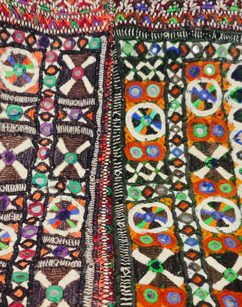 K&ouml;nnte beinhalten: Nahaufnahme eines farbenfrohen Textils mit aufwendiger Stickerei. Das Design zeigt geometrische Muster, darunter Quadrate, Kreuze und Kreise, in Orange-, Gr&uuml;n-, Blau- und Braunt&ouml;nen. Kleine, reflektierende Spiegel sind in das Design integriert und erzeugen einen schimmernden Effekt.