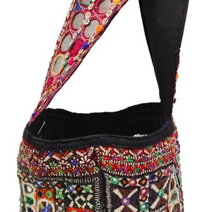 K&ouml;nnte beinhalten: Eine bestickte Umh&auml;ngetasche mit schwarzem Riemen. Die Tasche zeigt komplizierte Muster in Rot, Orange, Gr&uuml;n und Blau, mit kleinen Spiegeln und dekorativen N&auml;hten. Der Riemen ist mit Spiegeln und rotem Stoff verziert.