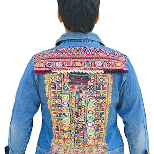 Op de afbeelding: Een spijkerjasje met levendige borduursels op de rug en manchetten. De borduursels tonen kleurrijke geometrische patronen met rode, gele, groene en blauwe draden. De jas heeft een klassieke kraag en knopen.