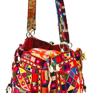 May include: A brightly coloured, handmade bag with a patchwork design. The bag features red, orange, yellow, and blue geometric patterns, decorated with shells and tassels. The straps are adorned with decorative stitching and metal rings.