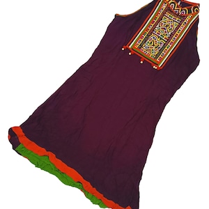 Puede incluir: Un vestido largo sin mangas en un tono burdeos oscuro. El escote presenta un panel bordado con patrones geométricos en rojo, amarillo, verde y blanco. El dobladillo está adornado con tela naranja y verde. Pequeñas borlas blancas adornan el escote.