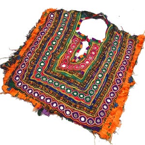 Puede incluir: Textil bordado colorido con escote cuadrado. La prenda presenta intrincados patrones en rojo, verde, azul y naranja, con pequeños detalles de espejo. Los bordes están adornados con flecos naranjas.