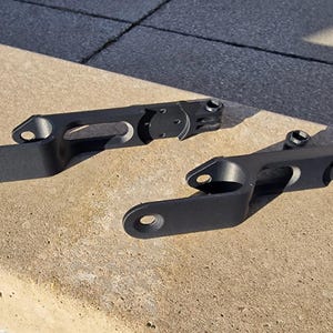 May include: Two matte black metal brackets with an elongated, angular design. Each bracket features a circular mounting hole at one end, a central rectangular cutout, and a hinged, circular attachment point at the opposite end with a small bolt.